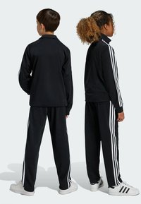 Černý sportovní teplákový komplet s bílými třípruhovými akcenty, s límcem na zip a volně střiženými kalhotami. Materiál: hladká tkanina.