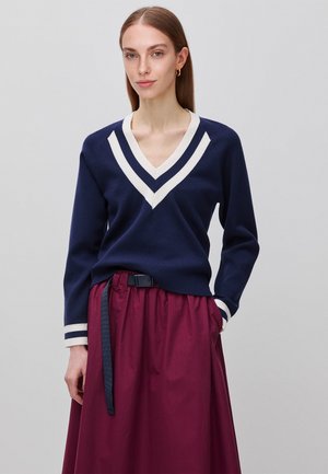 Vrouw draagt een marineblauwe V-hals trui met witte strepen, gecombineerd met een hooggetailleerde bordeauxrode rok met een zwarte riem.