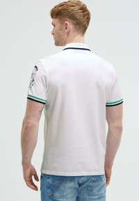 Weißes Polo-Shirt mit grünem und navyfarbenem geripptem Kragen, ausgestattet mit einem grafischen Design auf dem linken Ärmel und einer glatten Baumwolltextur.