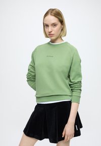 Sweatshirt verde de algodão com decote redondo e punhos canelados, combinada com saia plissada de bombazina preta com etiqueta lateral.