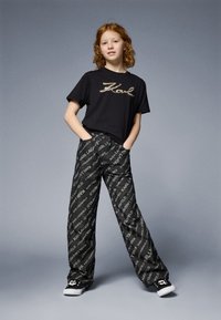 Μαύρο t-shirt με γκλίτερ λογότυπο "Karl"; χαλαρό, μοτίβο μαύρο παντελόνι με άσπρο κείμενο; φορώντας μαύρα παπούτσια με σχέδιο κρανίου.