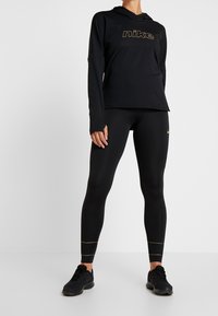 Felpa nera a maniche lunghe con logo Nike in oro, abbinata a leggings neri con dettagli dorati e scarpe da ginnastica nere con superficie strutturata.