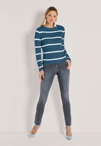 Frau steht vor einer hellen Wand und trägt einen blau-weißen gestreiften Pullover, graue Jeans, blaue High Heels und lange Perlenohrringe.