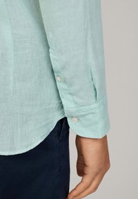 Camisa de lino color azul claro con puños abotonados, presentando un dobladillo ligeramente curvado y tejido de contraste en color navy por debajo. Acabado suave y texturizado.