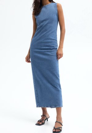 Woman wearing sleeveless blue denim midi dress and black strappy heeled sandals against white background.