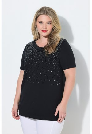 Vrouw met blond haar, gekleed in een zwart topje met korte mouwen met wit stippenpatroon en witte broek, staand tegen een witte achtergrond.