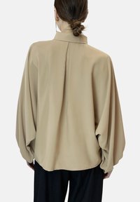 Losse beige blouse met een hoge kraag, geplooide achterdetails en volumineuze mouwen. Gemaakt van een soepele stof.