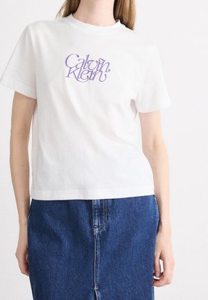 Femme portant un t-shirt blanc Calvin Klein avec un logo violet et une jupe en jean bleue sur fond blanc uni.