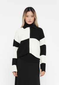 Cardigã de malha curto em preto e branco com fecho na frente, apresentando um design em blocos de cor e textura canelada. Mangas longas com gola.