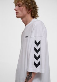 Fiatal személy göndör hajjal, aki fehér Hummel hosszú ujjú pólót visel, fekete chevron nyilakkal az ujjon és látható tetoválással a karján.