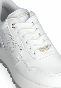 Zapatilla blanca de cuero con detalles perforados, cordones blancos planos y acento de logo en tono dorado, con una gruesa suela gris.