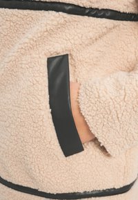 Main partiellement dans une poche d'une veste polaire beige clair texturée avec un liseré noir au bord de la poche et sur les coutures.