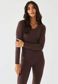 Brązowa dopasowana bluzka z długim rękawem i dekoltem w kształcie litery V oraz gładką fakturą, zestawiona z dopasowanymi spodniami o dopasowanym kroju, oba z elastycznego materiału.