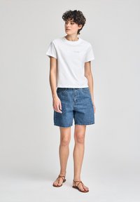 Vit t-shirt i bomull med logotyp, tillsammans med blå denimshorts. Sandaler med tunna remmar. Enfärgad bakgrund, fokus på kläddetaljer.
