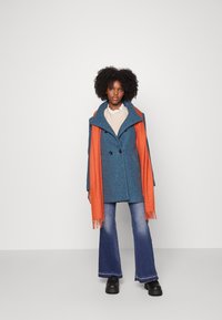 Manteau en mélange de laine bleu avec double boutons, col haut, et une étole orange avec des franges. Le mannequin porte un jean évasé et des chaussures noires.