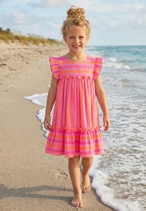 Smiling young girl with blonde hair in a pink and orange striped dress walking barefoot along the sandy beach near gentle waves.