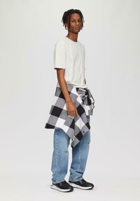 Witte t-shirt, lichtblauwe ripped jeans en zwarte sneakers. Zwart-wit geblokte flanelhemd om de taille geknoopt. Portretoriëntatie.