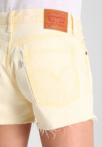 Pantalones cortos de mezclilla amarillos Levi's con dobladillo deshilachado, que cuentan con un bolsillo trasero y una etiqueta de marca beige con un logotipo gráfico. Material de algodón, diseño ceñido.