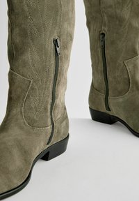 Botas de ante verde oliva hasta la rodilla con un sutil detalle de costura y cremallera lateral, que presentan un bajo tacón negro y una textura suave.
