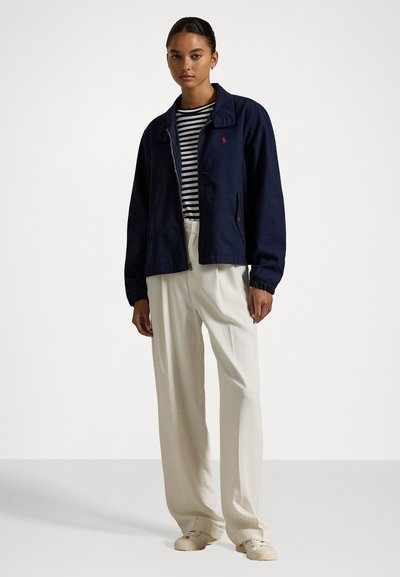 Navyblauwe, ritsjack met een puntige kraag, elastische manchetten en twee zakken; gedragen over een gestreept shirt en crèmekleurige, wijde broek.