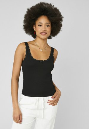 Femme avec afro portant un haut sans manches noir orné de dentelle, un pantalon blanc, un collier coloré, des boucles d'oreilles rouges et plusieurs bagues, debout avec la main sur la hanche.