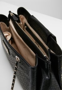 Sac fourre-tout noir avec un extérieur texturé embossé du logo, des détails en métal doré, et un intérieur beige clair comportant plusieurs compartiments et une fermeture éclair.