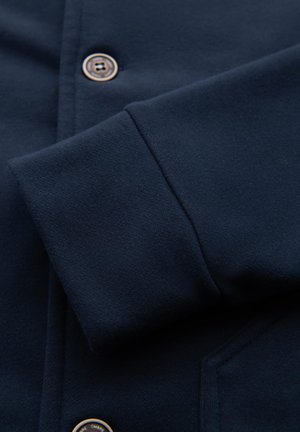 Marineblauwe button-up shirt met een textuurstof en een close-up van de manchette, met een metalen knop met een cirkelontwerp.