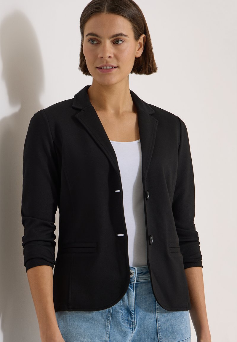 Femme aux cheveux bruns courts portant un blazer noir, un haut blanc et un jean bleu, debout contre un mur clair.