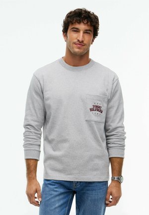 Mand iført lys grå langærmet skjorte med brystlomme tekst og blå jeans, stående mod en ensfarvet hvid baggrund.