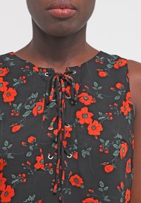 Schwarze Bluse mit floralen Motiven, die rote Blumen und grüne Blätter zeigt. Schnürkragen mit silbernen Ösen. Leichter, strukturierten Stoff.