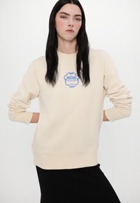 Felpa beige a girocollo con un logo grafico blu "Moschino Jeans Hotel" sul petto. Tessuto morbido, polsini e orlo a costine.