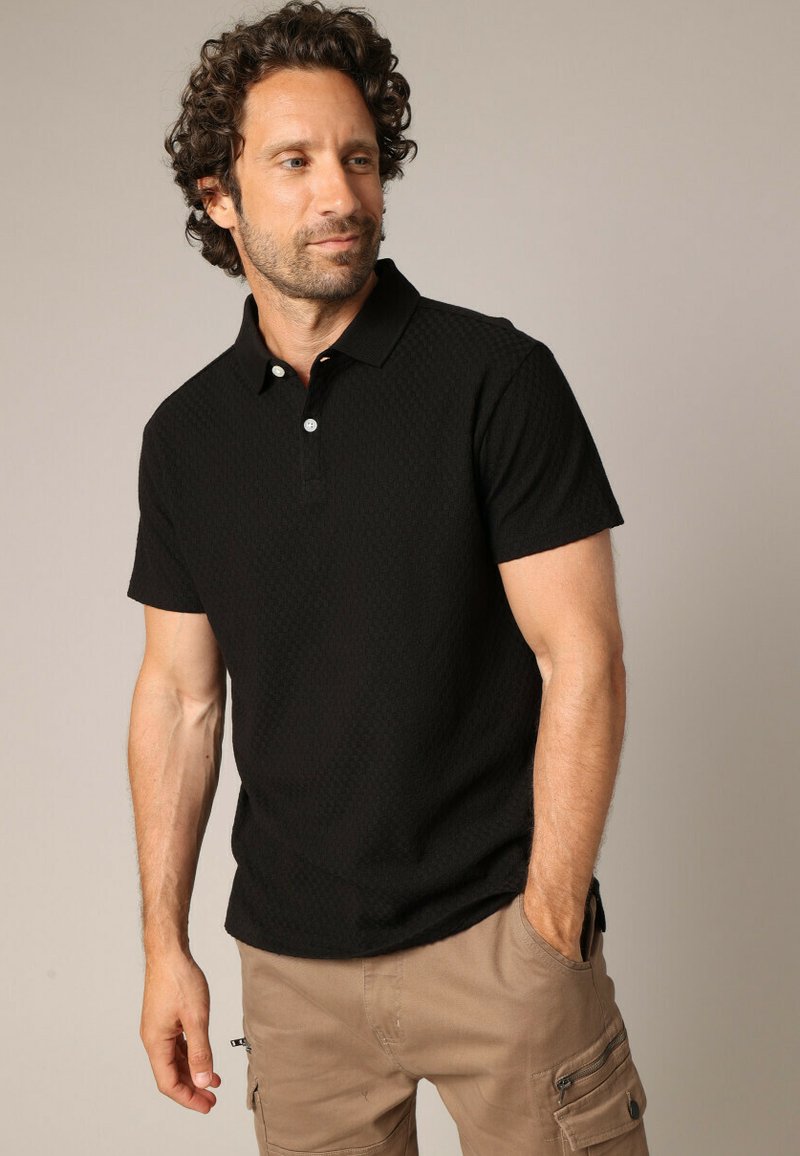 Polo-shirt noir à manches courtes avec un motif texturé, doté d'un col et d'un plastron à deux boutons. Porté avec un short cargo kaki.