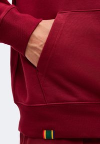 Main dans la poche avant d'un sweat-shirt rouge avec un bord-côtes et une petite étiquette en tissu colorée à l'ourlet.