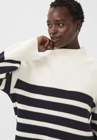 Gros plan sur une personne portant un pull à col montant côtelé avec de larges rayures horizontales noires et blanches, touchant son cou d'une main.