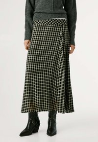 Lunga gonna a scacchi bianca e nera in tessuto morbido, con design drappeggiato, indossata con un maglione grigio e stivali neri con tacco.