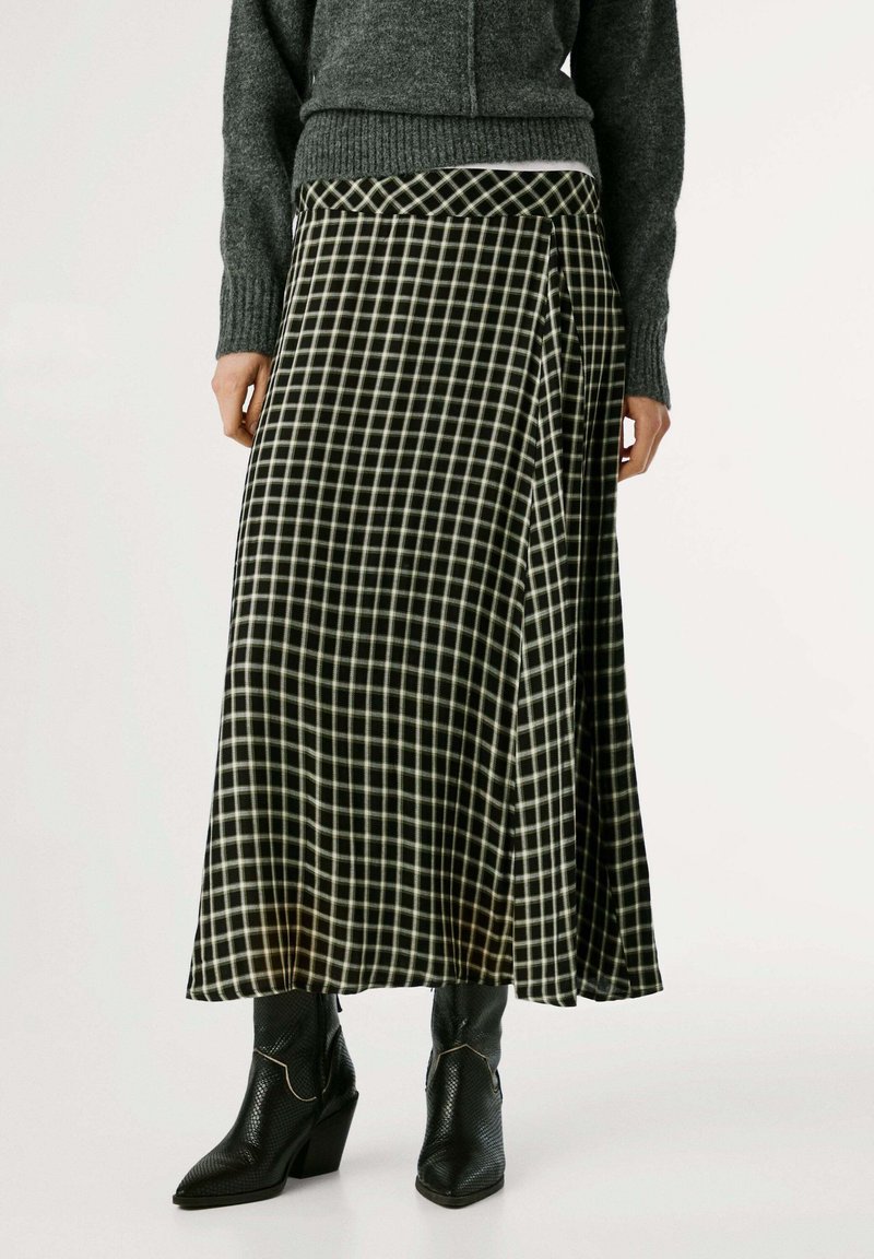Lunga gonna a scacchi bianca e nera in tessuto morbido, con design drappeggiato, indossata con un maglione grigio e stivali neri con tacco.