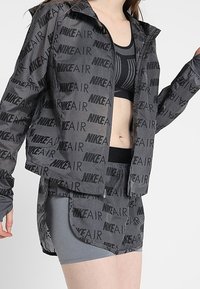Ensemble de vêtements de sport gris comprenant une veste à zip avec un imprimé répété "NIKE AIR", un design cropped, et une jupe ajustée assortie.