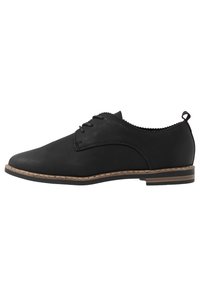 Chaussure en cuir noir avec une finition lisse, bout rond et couture décorative. Présente un talon bas et une semelle texturée avec une bordure beige contrastante.