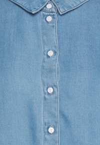Camisa de denim azul claro com colarinho pontudo, apresentando uma pala de botão com botões brancos e uma textura suave.