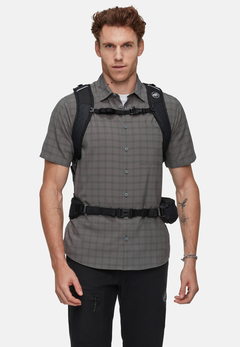 Kortermet grå rutete skjorte med knapper og krage, brukt under et svart sele med justerbare stropper og spenne foran.