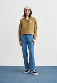 Καφέ φλις πουλόβερ με φερμουάρ μέχρι τη μέση και ταμπέλα με λογότυπο, συνδυασμένο με μπλε τζιν παντελόνι με φαρδιές παντελόνες και μαύρα αθλητικά παπούτσια με λευκές λεπτομέρειες.