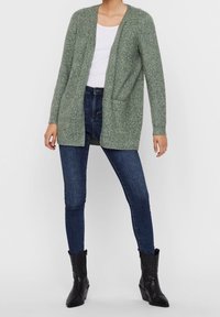 Knitted green cardigan, open front, textured fabric, two front pockets. Paired with dark blue skinny jeans and black heeled ankle boots.