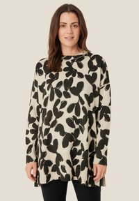 Sweter dzianinowy w kolorze kremowym z dużym czarnym wzorem liścia. Długie rękawy, okrągły dekolt, luźny krój, teksturowe prążkowanie u dołu i przy mankietach. Czarne spodnie.