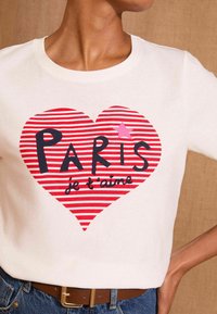 Balta marškinėliai su dideliu raudonai balto dryžuoto širdies grafinu, kuriame yra užrašas "PARIS" didelėmis juodomis raidėmis ir "je t'aime" rašomuoju šriftu.