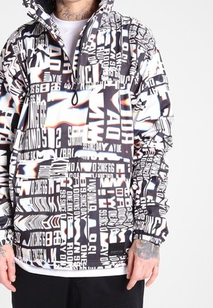 Homme portant un pull à manches longues noir et blanc avec texte mixte audacieux et motifs graphiques, fermeture éclair à l'avant, et poignets élastiques.