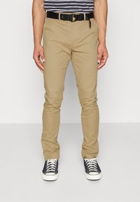 Chino khaki con vestibilità slim, realizzati in cotone leggero, dotati di una vita standard e tasche a fessura, abbinati a sneaker nere.