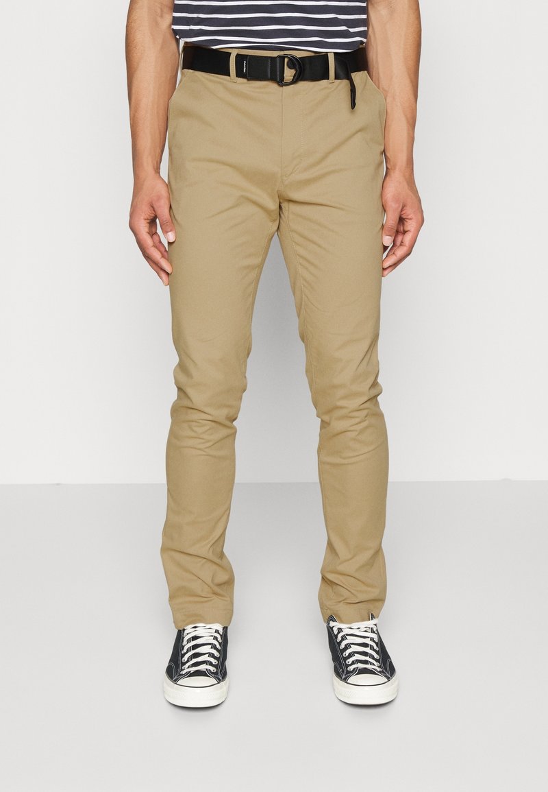 Chino khaki con vestibilità slim, realizzati in cotone leggero, dotati di una vita standard e tasche a fessura, abbinati a sneaker nere.