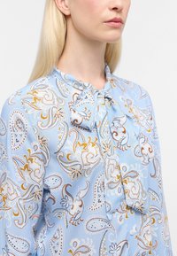 Blusa azzurro chiaro con motivo paisley in oro e bianco. Presenta un dettaglio con nodo al collo e una texture di tessuto morbida e liscia.