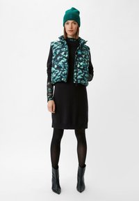 Schwarzes Kleid mit geripptem Saum, schichtweise getragen mit einer pufferweste in Teal und Schwarz mit Muster. Schwarze Strumpfhosen und spitze schwarze Ankle Boots runden den Look ab.