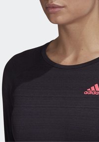 Musta urheilullinen pitkähihainen paita, jossa on teksturoitu kangas, vasemmassa rinnassa vaaleanpunainen Adidas-logo ja pyöreä kaulus.